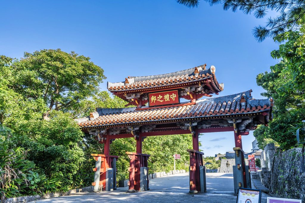 Shureimon Gate at Shuri Castle, a red lacquered wooden gate with a tiled roof and a plaque reading Shurei no kuni, framed by trees under a clear blue sky.