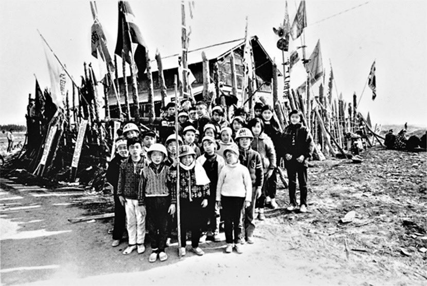 Children, wearing riot helmets stand at attention in front of a fortified hut. These are members of the Boys Corps of the anti-Narita Airport Hantai Domei.