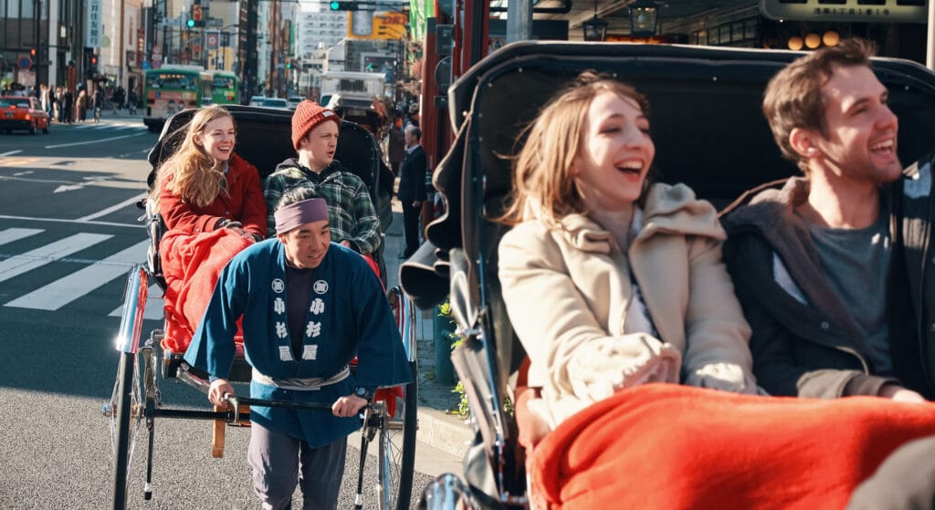 Two pairs of international tourists ride rickshaws in Japan.