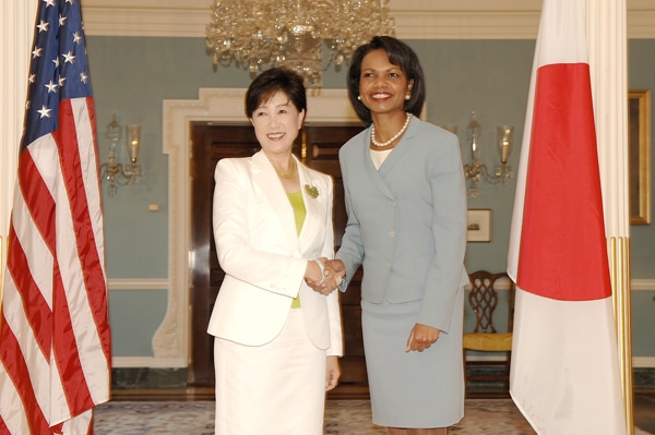 Koike Yuriko in a white outfit shakes hands with Condoleezza Rice, in a grey-blue outfit. On one side is the American flag, on the other, the Japanese flag.