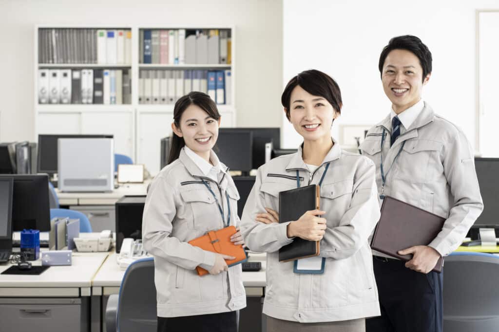 A Japanese workplace, with two women and one man in grey uniforms standing in front of desks and cabinets.