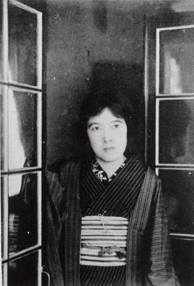 Yosano Akiko, a Japanese woman in kimono, stands behind a window in a black and white photograph. She did not see "mama" and "papa" as problematic.