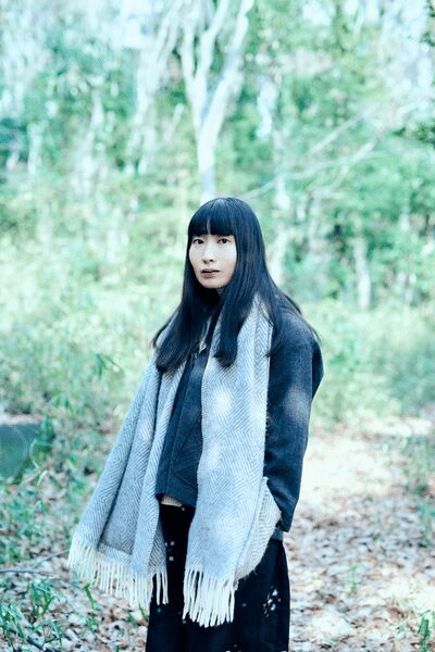 Saho Terao stands in front of forest wearing a long grey scarf and grey jacket.