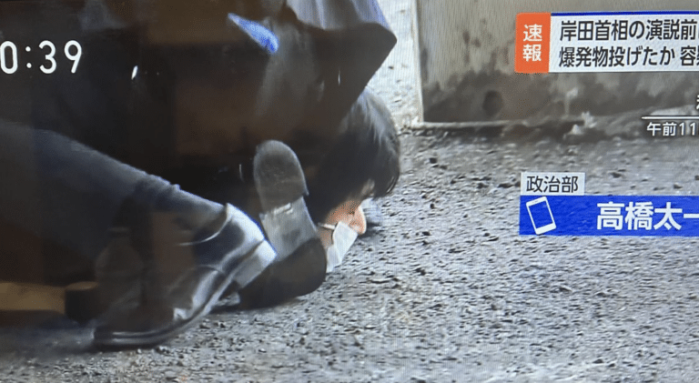 Screenshot shows a young man with black hair and white mask tackled after attempting assassination on Prime Minister Kishida.