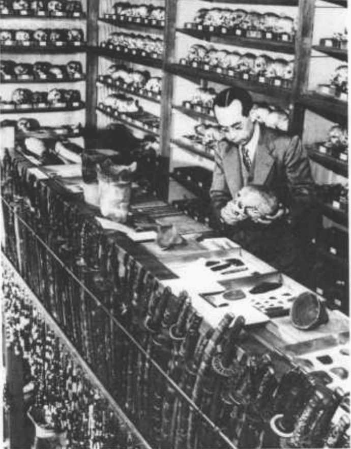 In a black and white photograph, a Japanese man in a suit stands amongst numerous Ainu remains and artifacts, including numbered skulls and countless swords.