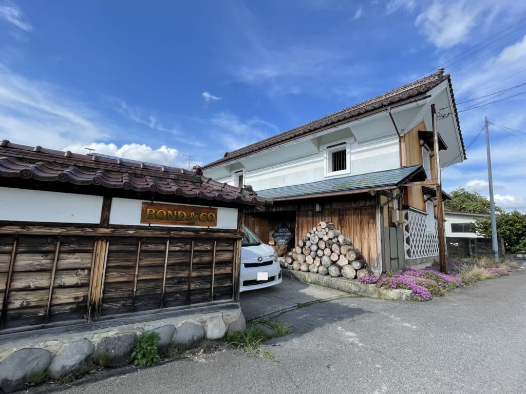 Wooden kura warehouse with sloping Japanese-style roof, wood pile, and wall with sign reading "Bond&Co." carved out of painted wood.