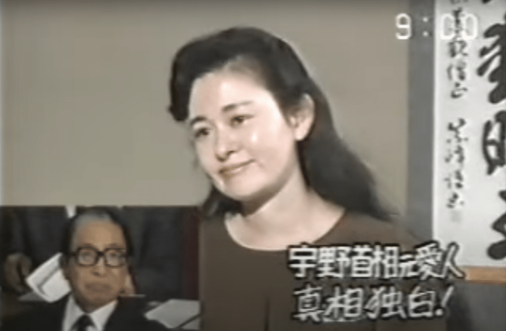 Former geisha Nakanishi Mitsuko in a simple brown shirt, appearing on TV channel TBS. A scroll bearing kanji is on the wall behind her, and a picture of Prime Minister Uno Sosuke is superimposed.