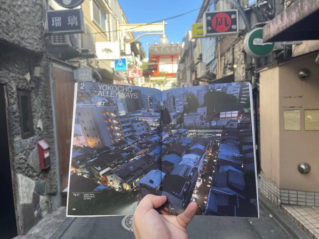 The book Emergent Tokyo open to a double page image of the densely packed bars of Golden Gai, while behind the book is an actual Golden Gai alleyway.