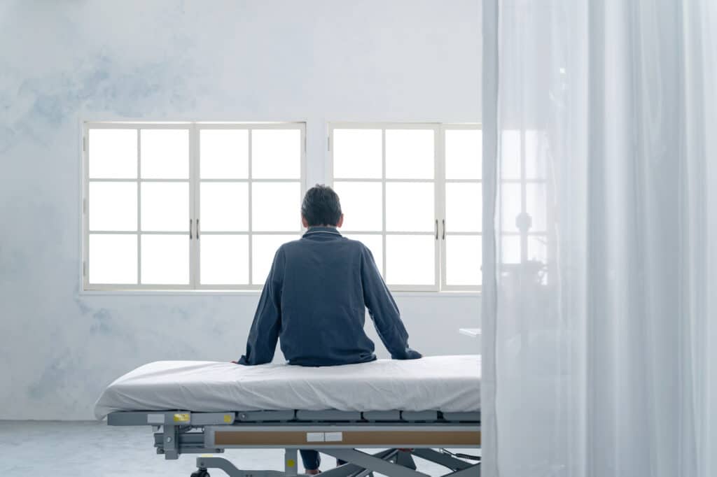 An individual sits on a hospital gurney with their back to the viewer, looking out two bright windows.