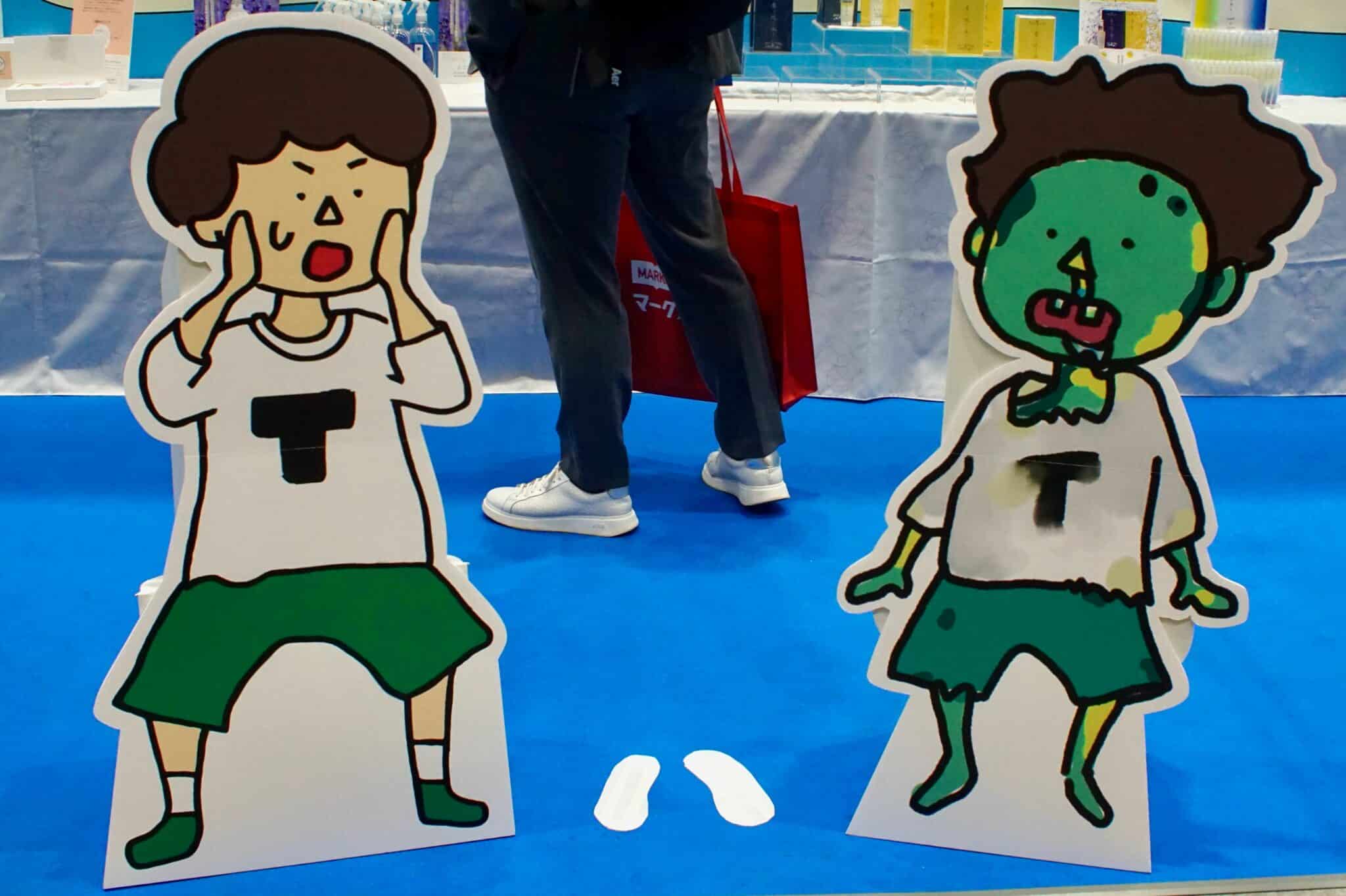 Standees of a cartoon boy in a white t-shirt with a "T" logo as he puts his hands to his face in shock upon seeing a standee of himself in a state of decomposition, looking like a cartoon zombie, at the showroom floor at ENDEX.