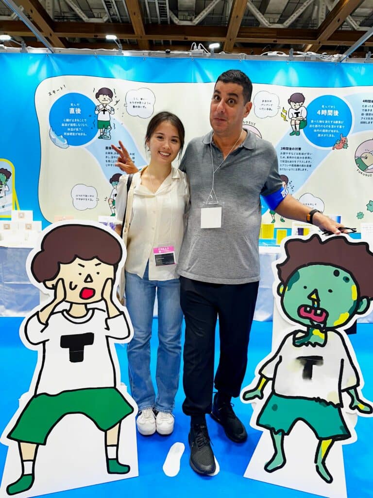 Jake Adelstein and Amy Yoshida-Plambeck stand between two cartoon standees of a boy shocked to see himself in a state of decomposition on the showroom floor of ENDEX.