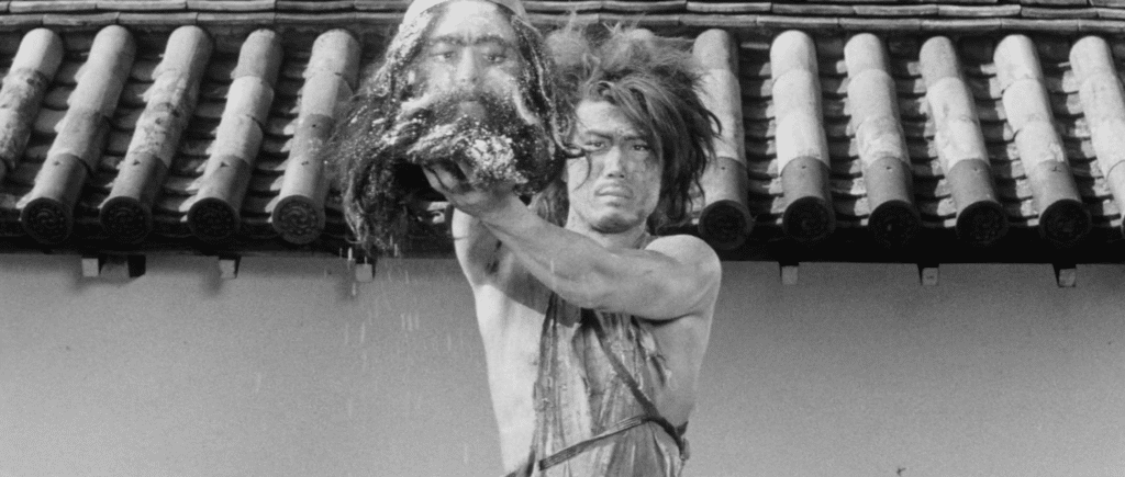 A ragged man with long hair stands in front of a Kyoto wall and lifts up the detached head, clearly a dummy, of a man with a long beard in the film Kuroneko.