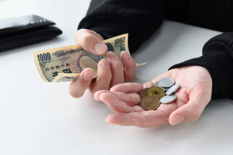 A person holds onto Japanese yen bills and spare coins, their gestures appearing upset. An empty wallet lays on the table nearby.