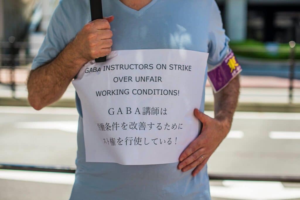 Protesting man in blue shirt and union badge with sign in English and Japanese saying "Gaba instructors on strike over unfair working conditions!"