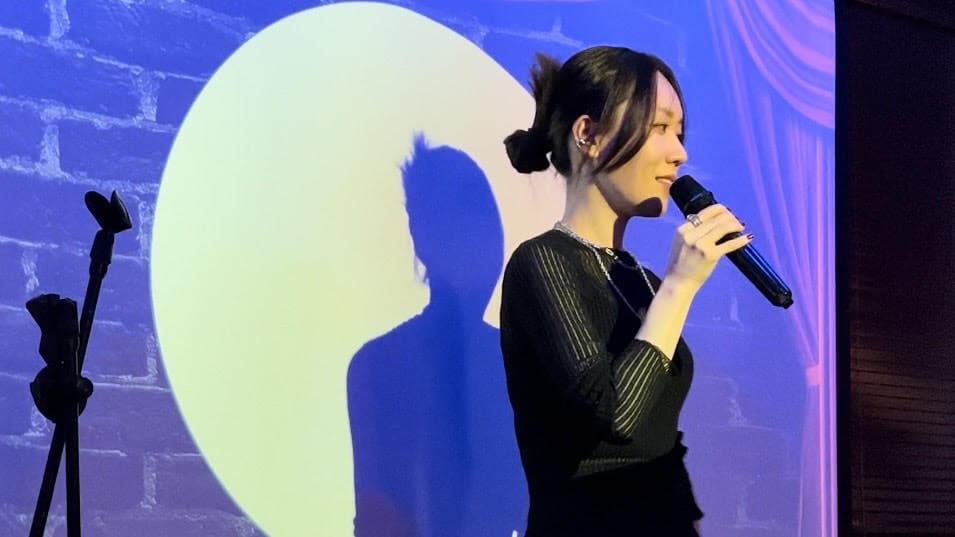 Comedian Yuki Nivez speaks into a mic while a spotlight illuminates her in front of a projected brick wall.