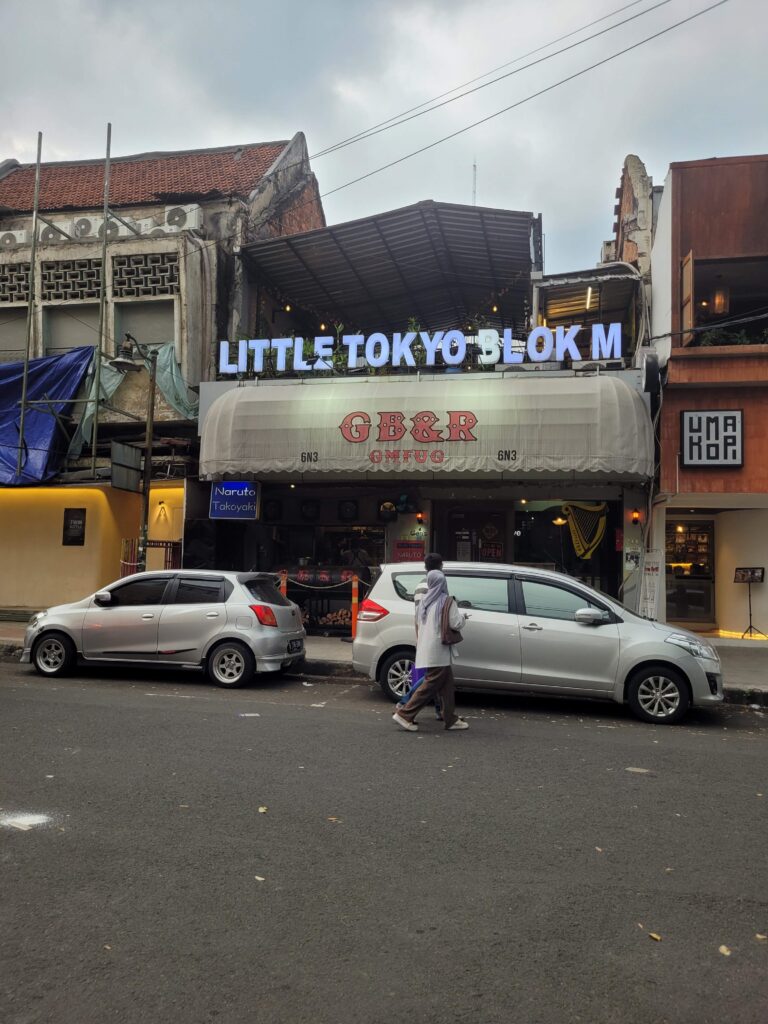 A woman an man walk on public streets in front of storefronts, with a large sign baring the words Little Tokyo Block M in florescent letters on the building fornt.