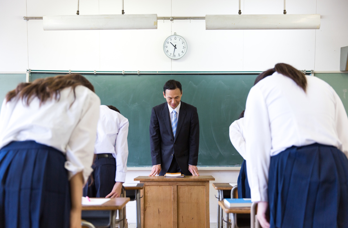 Picture of two aisles of students bowing to a teacher. The picture is taken from behind the students and at the end of the row, you can see a male teacher standing behind a podium and making his own small bow.