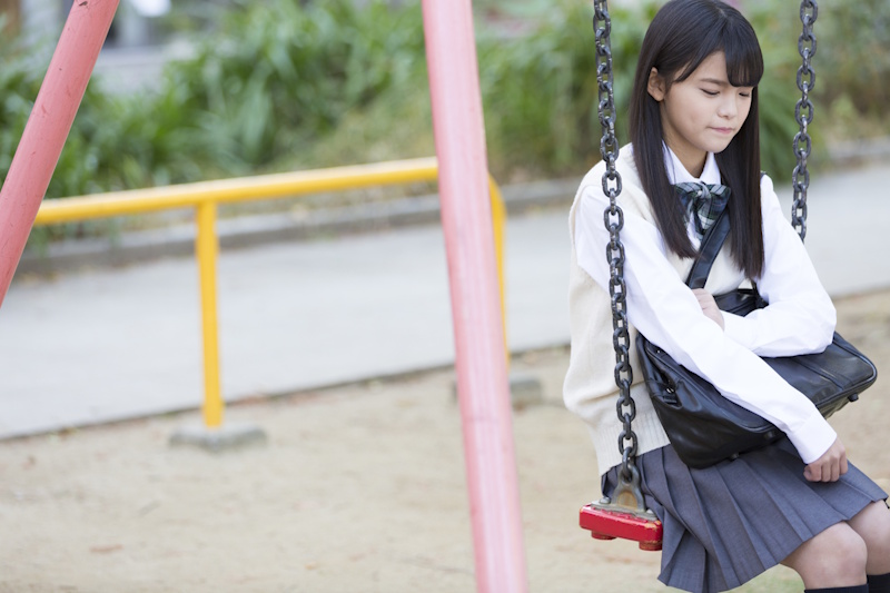 Picture of schoolgirl in school uniform sitting on a red swing on the right side of the screen, holding a shoulder bag in her lap and looking forlorn
