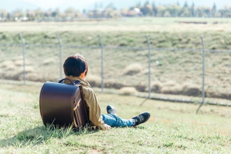 Kid with a randoseru style Japanese backpack sitting in a field with his back to the camera and looking very depressed