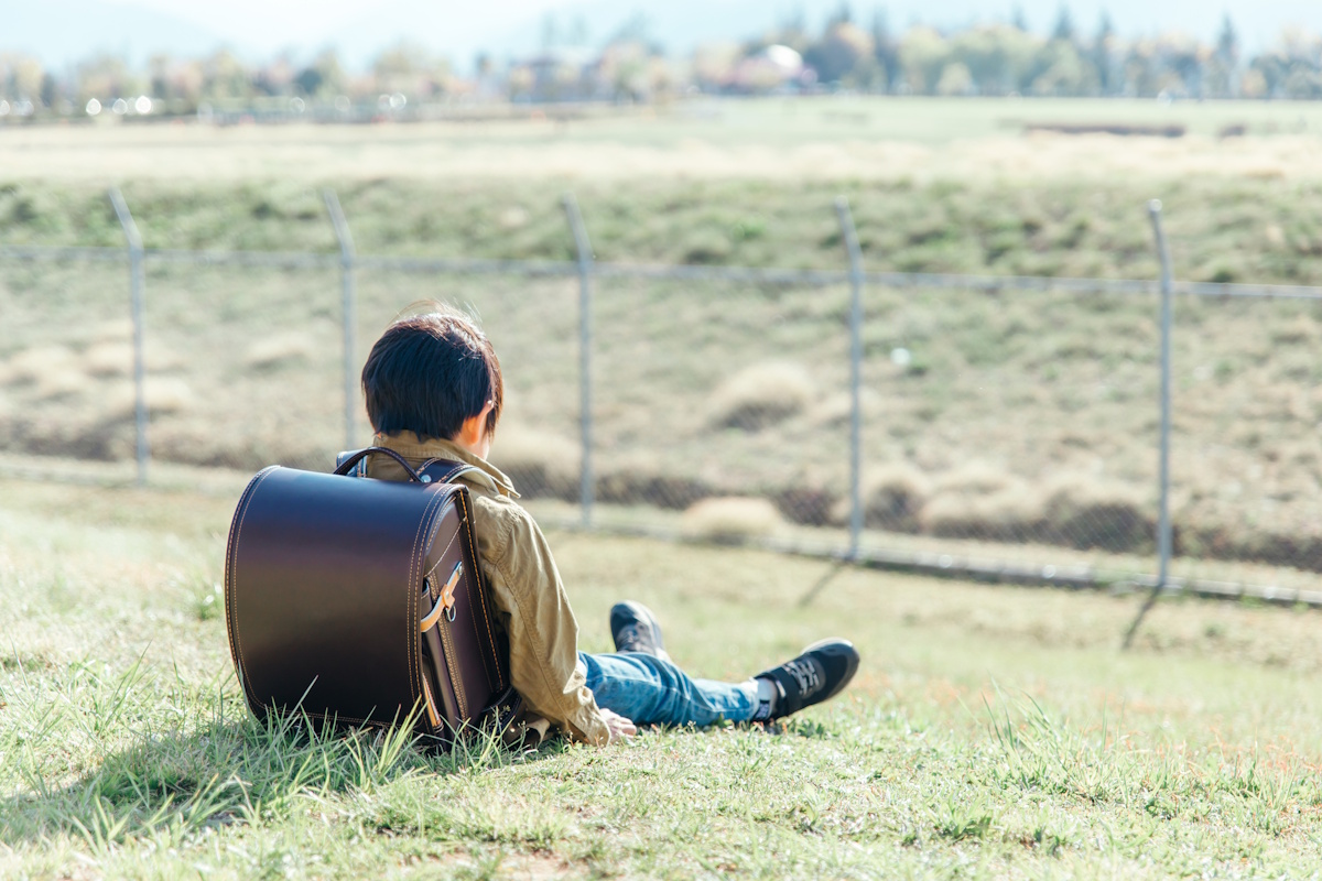 Kid with a randoseru style Japanese backpack sitting in a field with his back to the camera and looking very depressed