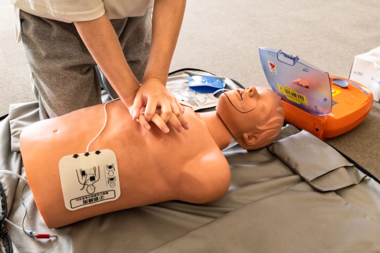 Person delivering CPR on a resuscitation dummy, with an AED device attached via pads and the device itself set above the dummy's head to the right
