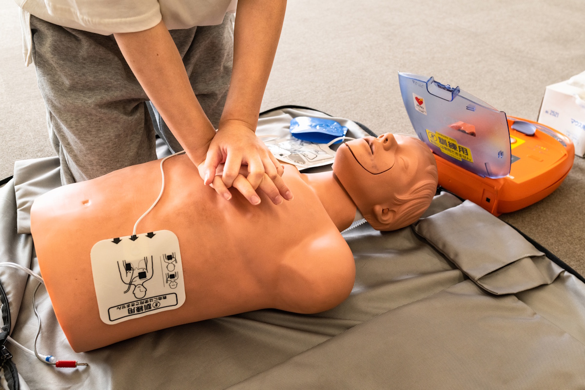 Person delivering CPR on a resuscitation dummy, with an AED device attached via pads and the device itself set above the dummy's head to the right