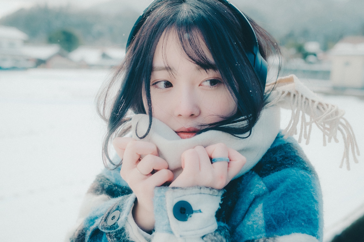 Young woman listening to mislc in the snow wearing a heavy blue winter coat and a tan scarf, with her hands held up to her chin while she looks off-camera to the right