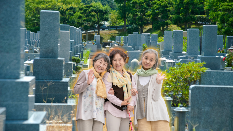 Grave friends story - picture of three older women with arms interlocked and smiling and waving to the camera. Behind them is a cemetery.