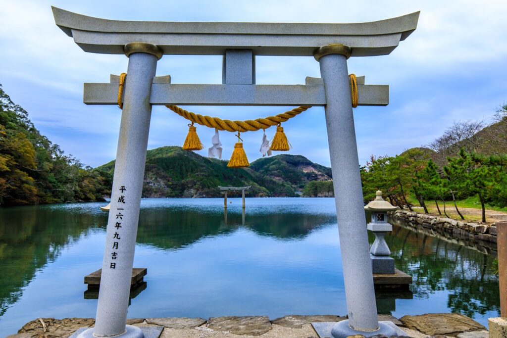 Picture of a white Shinto torii gate facing a lake of water. Watadzumi Shrine, Nagasaki Prefecture