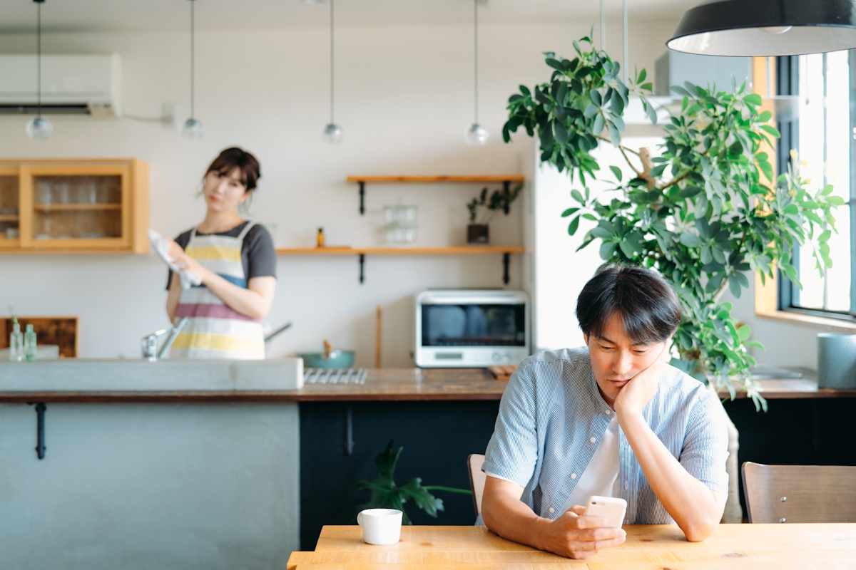 Wife washing ditches looks scornfully at husband sitting at kitchen table and playing on his smartphone