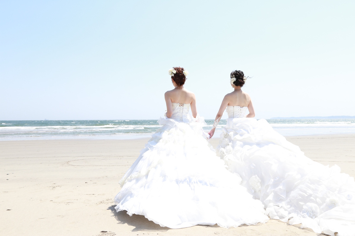 Two women in white wedding dresses walking towards the ocean on the beach, their backs to the cameras