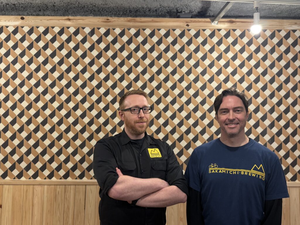 Matt and Daniel of Sakamichi Brewing stand in front of a wood-paneled walk with bare ceiling in their new Sakamichi North location.