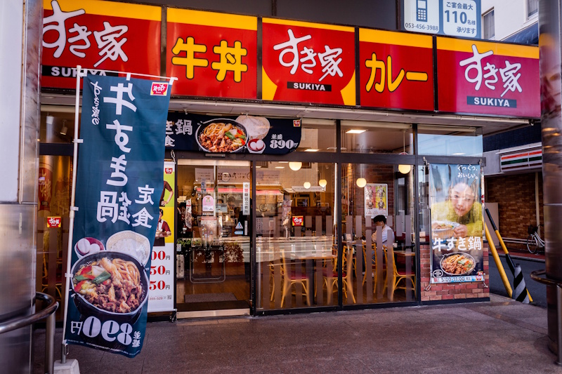 Picture of a Sukiya storefront.  Red sign above the stor with lettering in white and orange yellow. A banner on the left advertises its beef tendon bowl