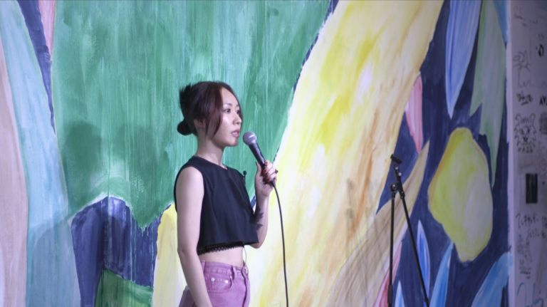 Yuki Nivez, microphone in hand, stands in front of a brightly colored wall.