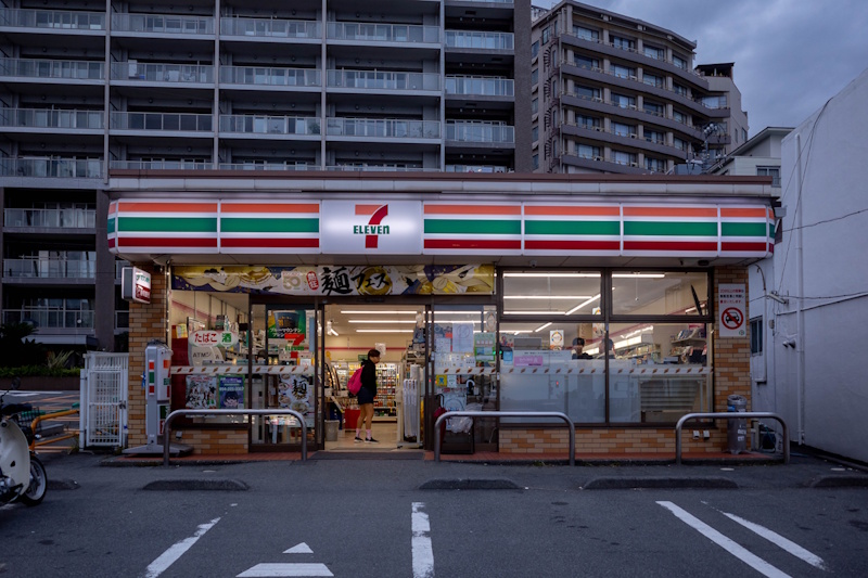 Picture of a 7-11 late at night in Japan from  the outside with the door open. A man with a red backpack is standing by the register.