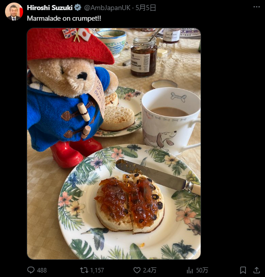 X post from @AmbJapanUK. Shows a Paddington bear doll with a crumpet. Text: "Marmalade on crumpet!"