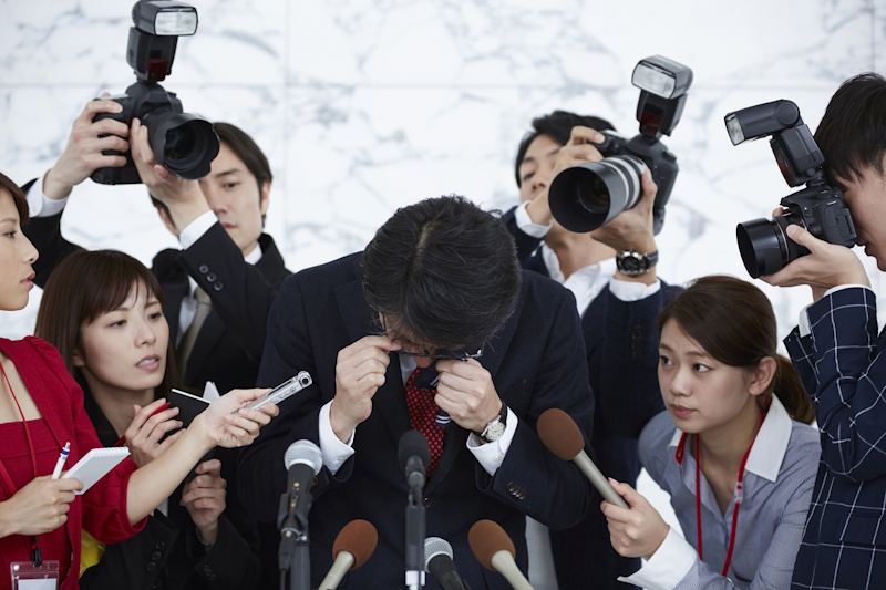 Man in suit bows in apology, holds glasses, as reporters take pictures and record his comments