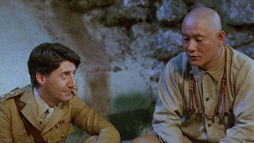 A film still from Merry Christmas, Mr. Lawrence, with Takeshi Kitano with a bald head in khaki and wearing prayer beads next to a British officer in uniform.