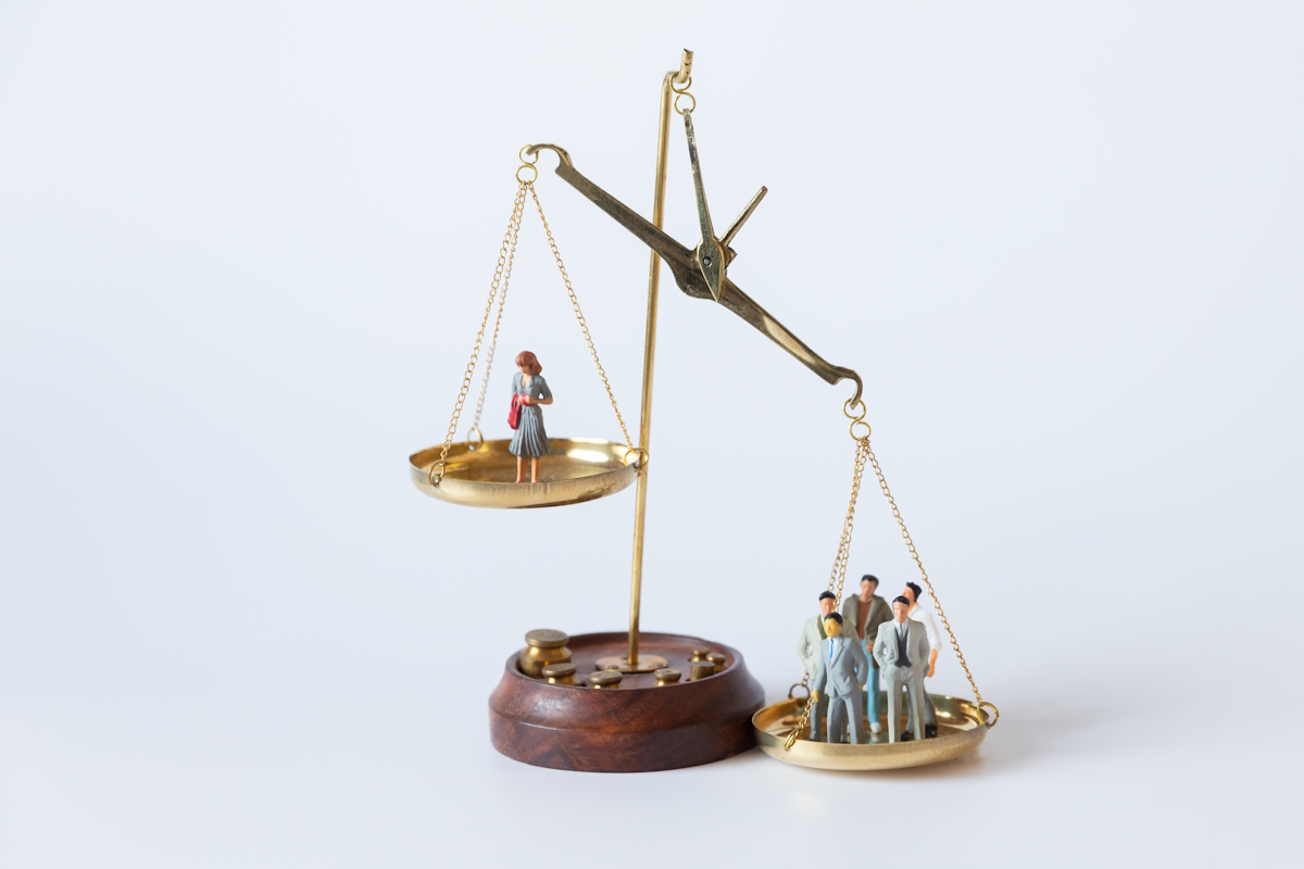 A set of scales with a single woman on the left being outweighed by a group of men on the right. The people are represented using dolls.