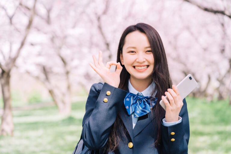 Woman with a phone in front of sakura trees holding up an OK sign with her fingers