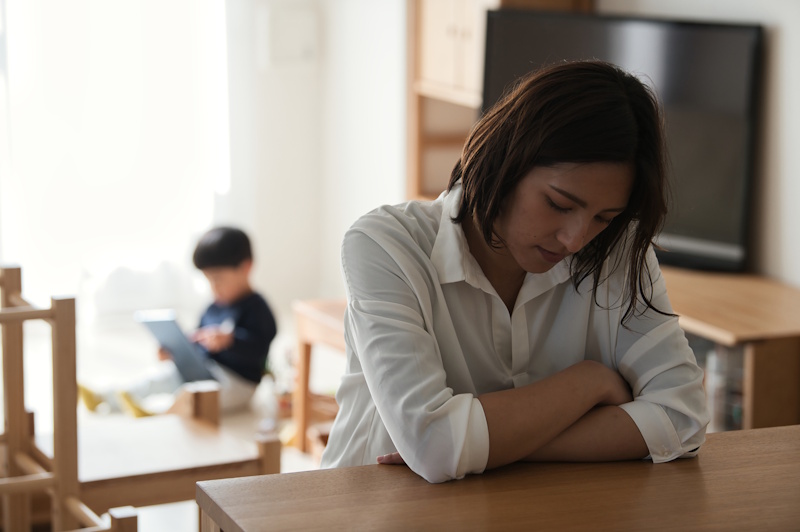 A woman with her hands folded on a table and looking like she's about to fall asleep. In the background, a young boy plays on a tablet