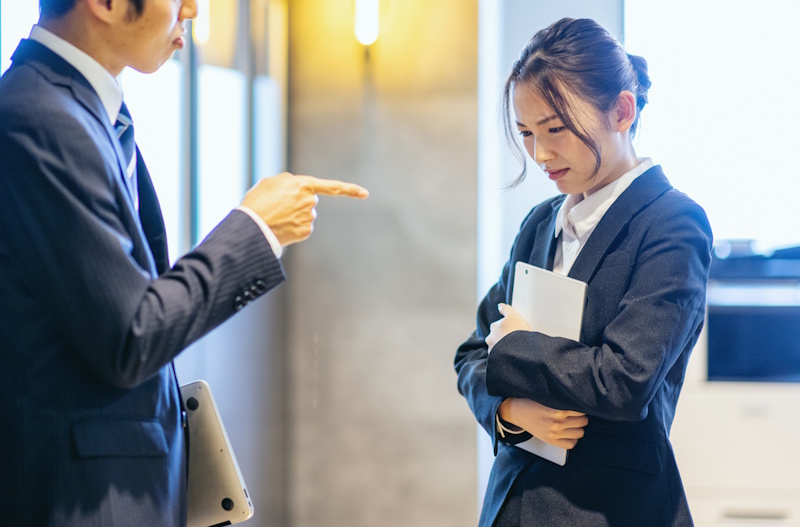 A male office worker pointing a finger at a female office worker. He's scowling while she looked cowed.