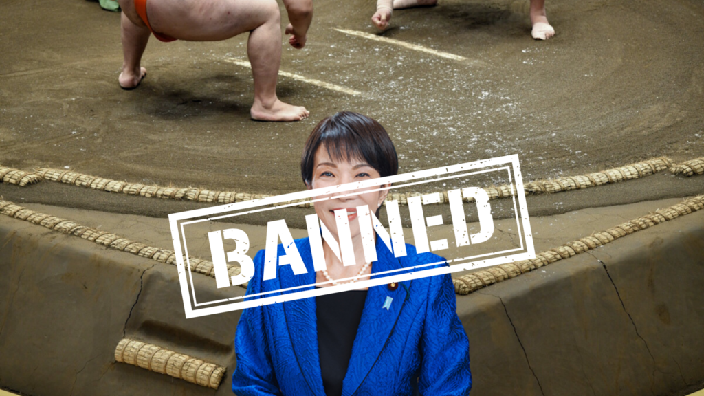 Two wrestlers facing off in the sumo ring behind a picture of Takaichi Sanae showing her banned from the ring