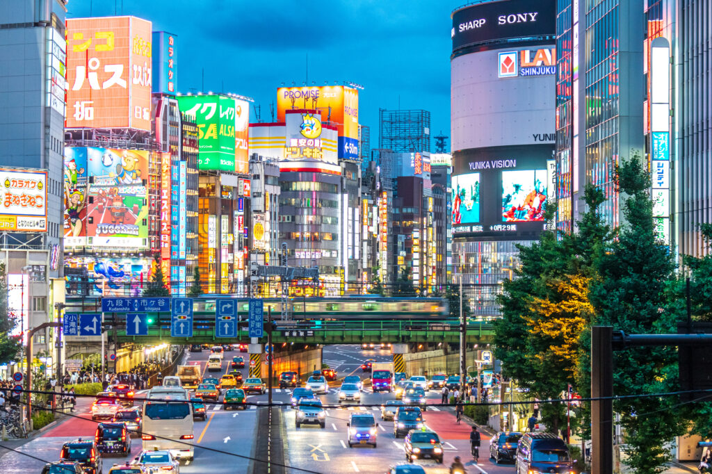 Yasukuni Avenue in Shinjuku bustles with car and is illuminated by hundreds of signs and advertisements. An elevated Yamanote Line train speeds by.