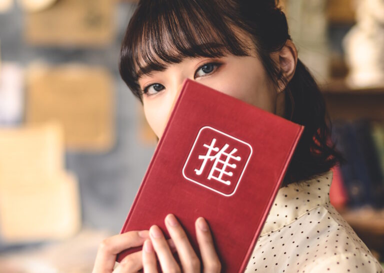 Woman holding up a red book with 推 (the kanij for oshi) in front of her face