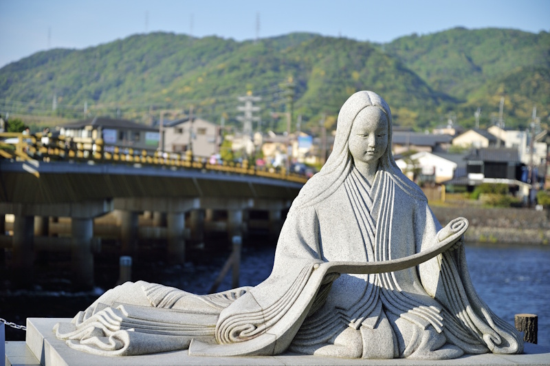 Stone statue of Heian-era author Murasaki Shikibu seated in flowing robes near Uji Bridge, with the river and hills behind