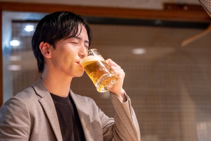 A man in a suit jacket drinks a beer from a glass mug at a bar.