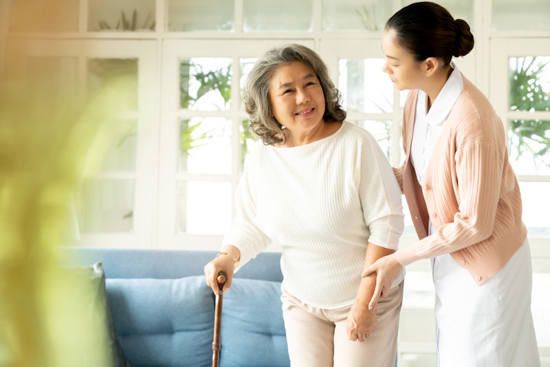 A young caregiver supports an elderly woman with a cane in a bright living room.