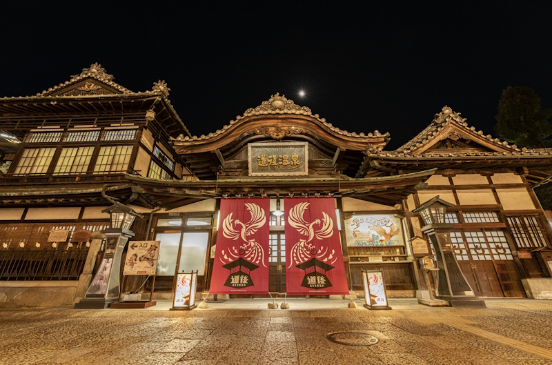 Night view of the Dogo Onsen main bathhouse in Matsuyama, a wooden structure with red phoenix-patterned curtains at the entrance.
