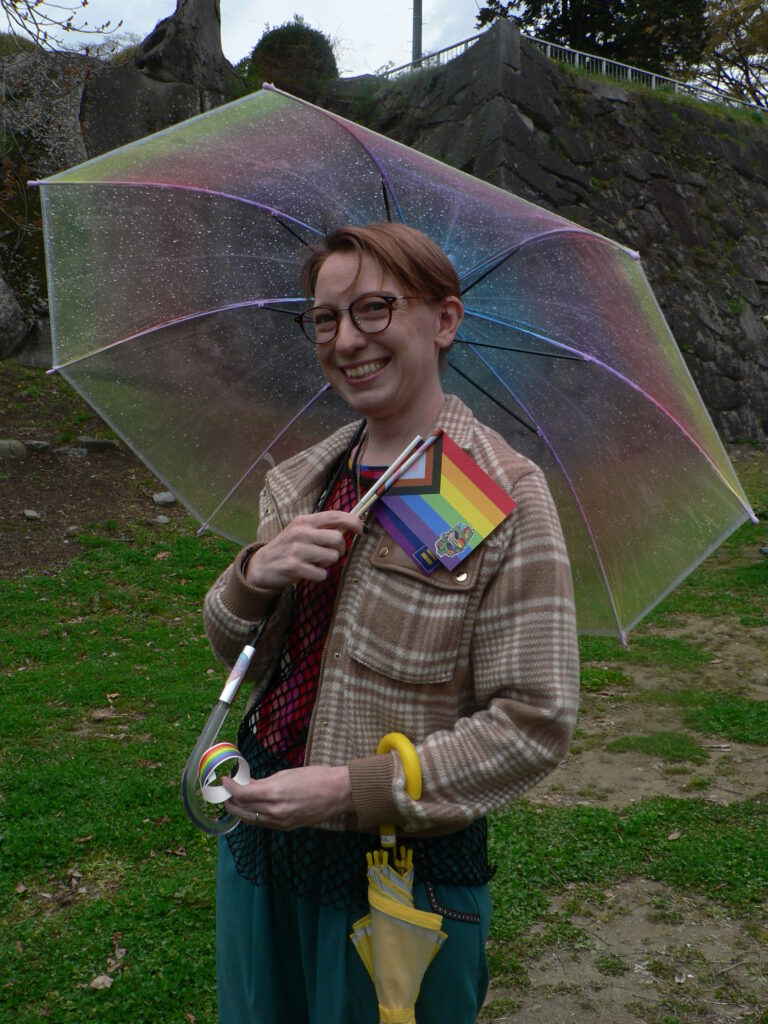 Chris Takisawa smiles while holding a rainbow umbrella and Progress Pride flag at Morioka Castle Ruins Park